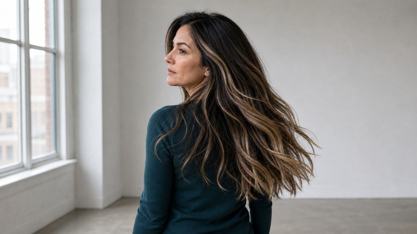 A woman turns beside a window, showing a retained dark brunette root, staggered neutral-beige balayage ribbons, and brighter wavy ends