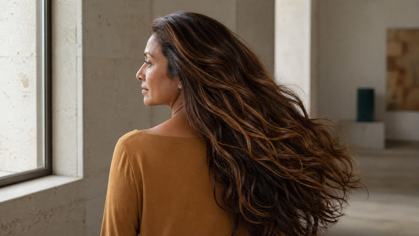 Woman in profile turning beside a window, with long medium-deep chestnut waves, brown-copper mids, and narrow deep-golden ribbons over a brunette root